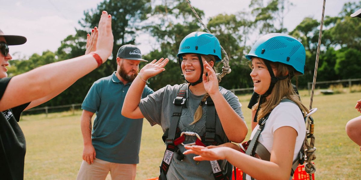 two girls at holiday camp in Melbourne wearing helmets and giving high fives after completing adventure activities.