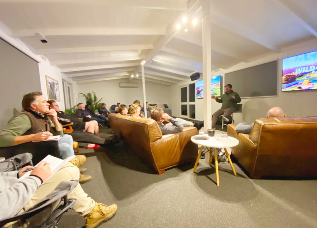 Image of facilitator training event with instructor speaking to people sitting on couches in conference room with visual and audio screens for viewing.